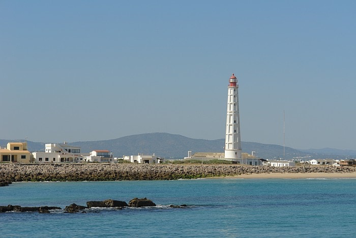 A lighthouse and small buildings sit on a rocky island outcrop surrounded by calm, blue-green water.