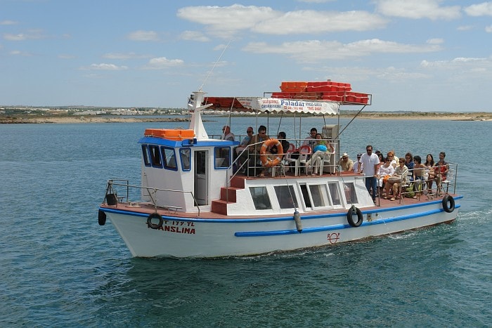 A small passenger ferry with a blue and white hull motors through calm turquoise water near a low-lying island.