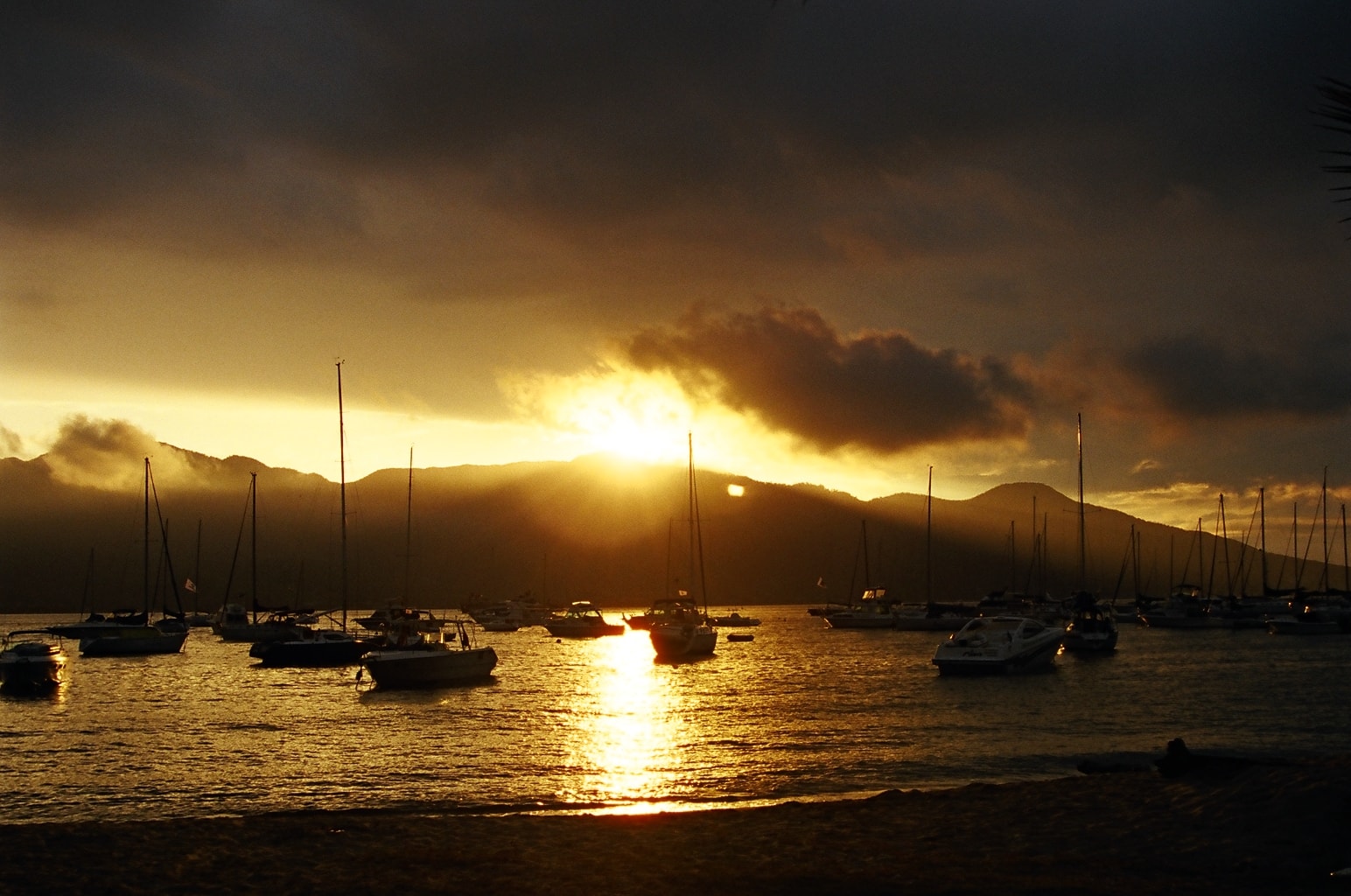 A dramatic sunset over the water in Ilhabela, with sailboats anchored in the harbor and mountains silhouetted against a colorful sky.