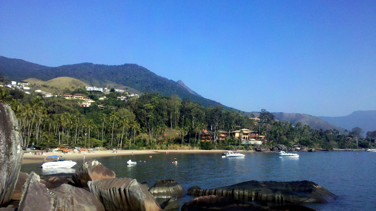 A wide view of an inlet on Ilhabela, showing calm waters, palm trees, boats, and the forested mountains rising behind the shoreline.