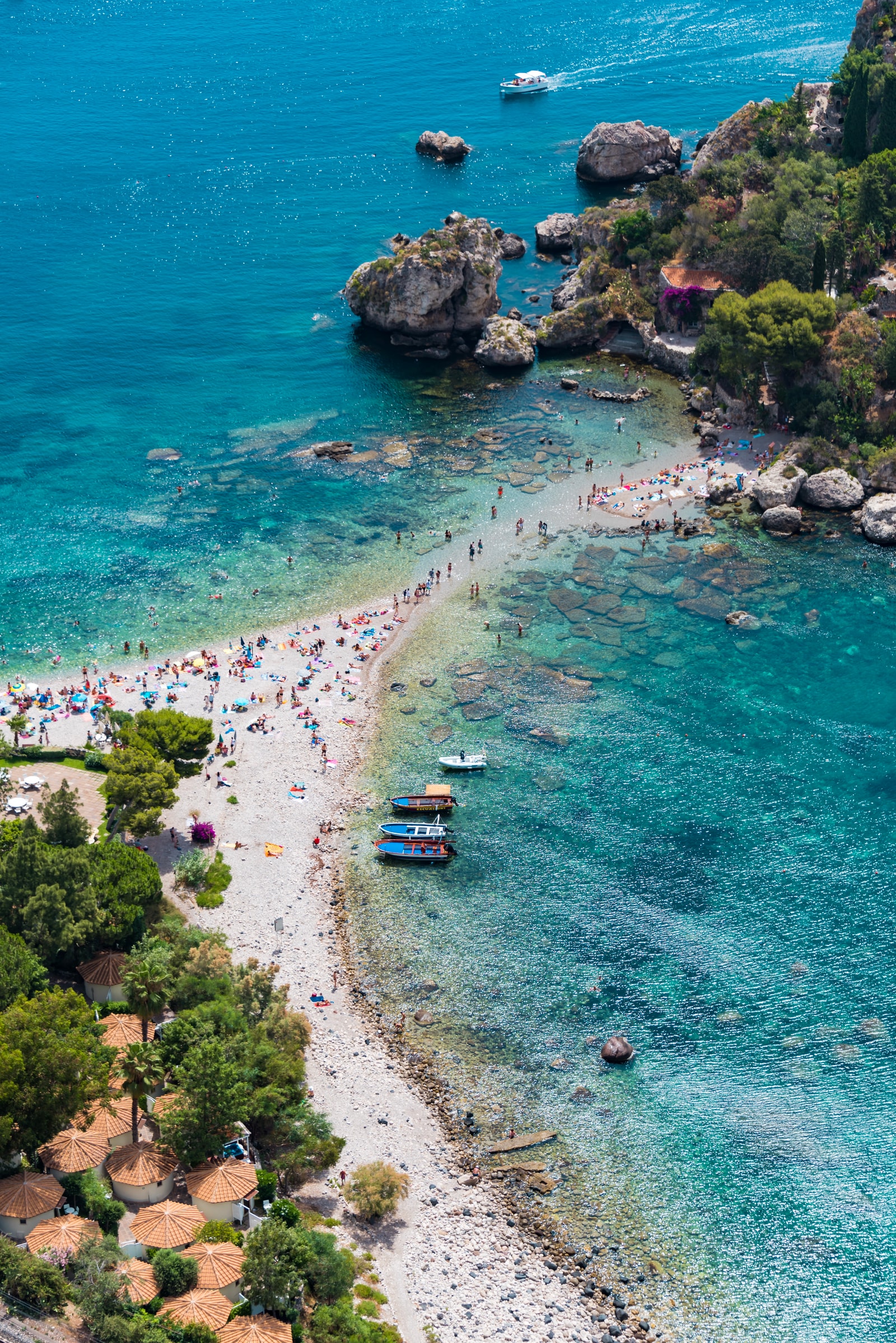 A vertical aerial travel shot of the thin pebbled sand path connecting the Sicilian mainland to Isola Bella reserve, with crystal clear turquoise waters.