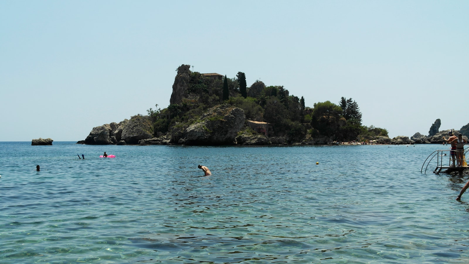 The lush, green islet of Isola Bella rises from the crystal-clear, shallow turquoise waters of the Ionian Sea, with a historic stone house visible among the trees.