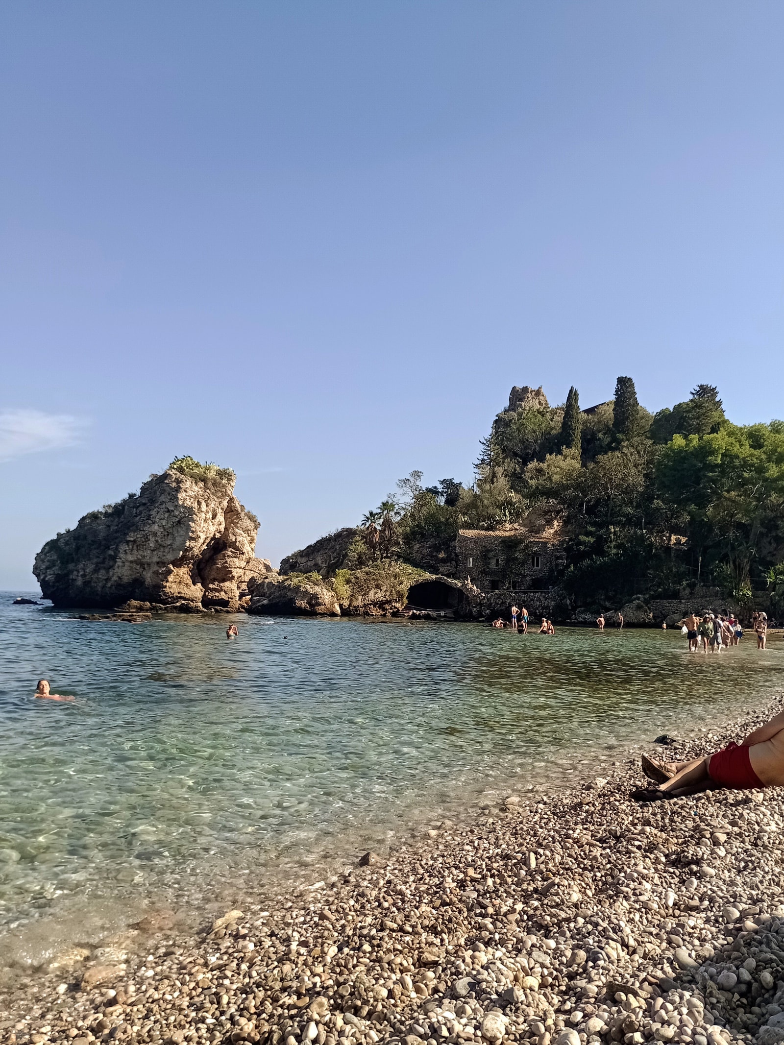 Beachgoers wade through the shallow water on the pebble beach of Isola Bella, with historic fortress ruins visible on the hills above.