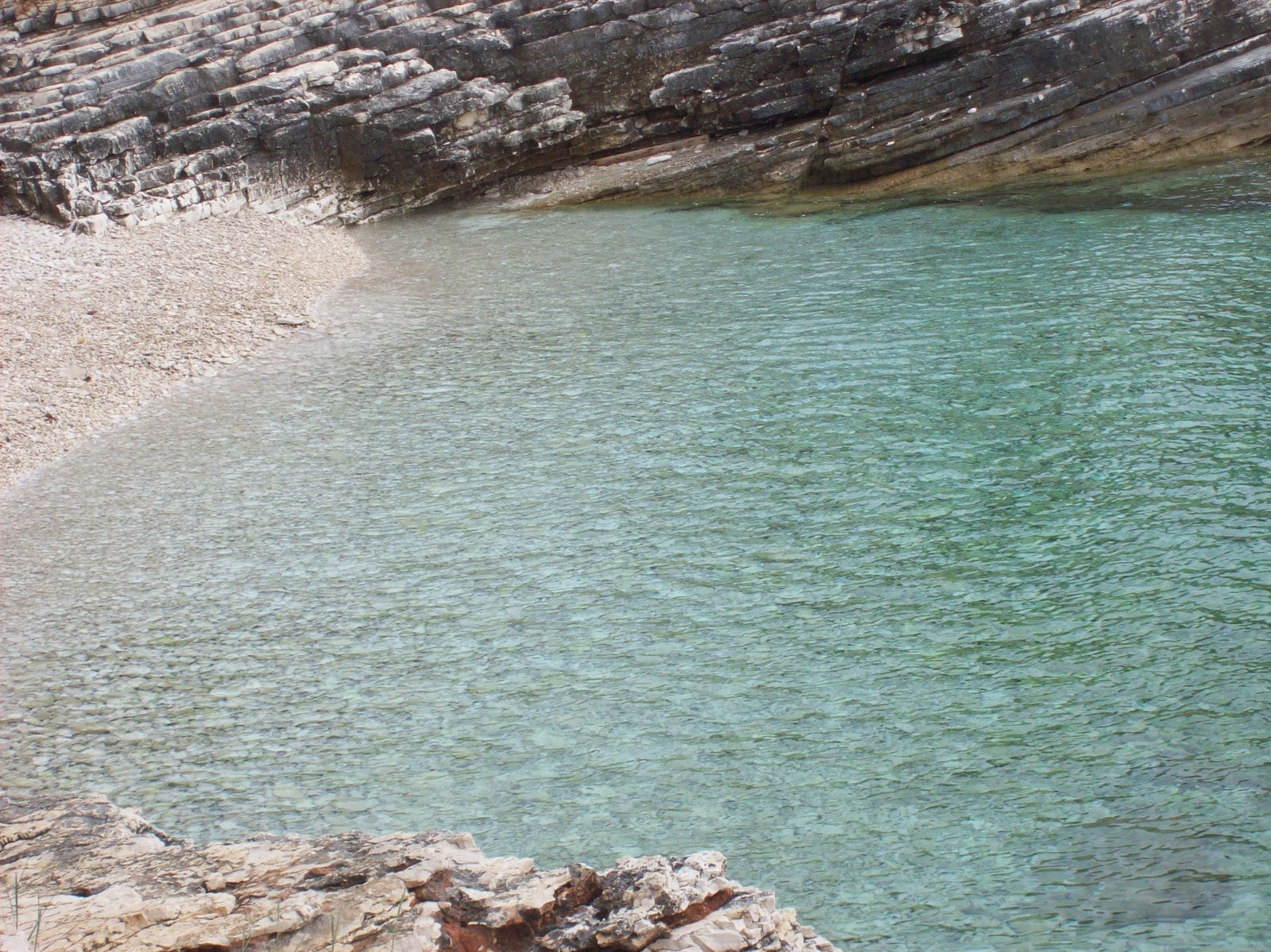 A secluded cove with incredibly clear, turquoise water is surrounded by rugged, rocky shores and green Mediterranean vegetation.