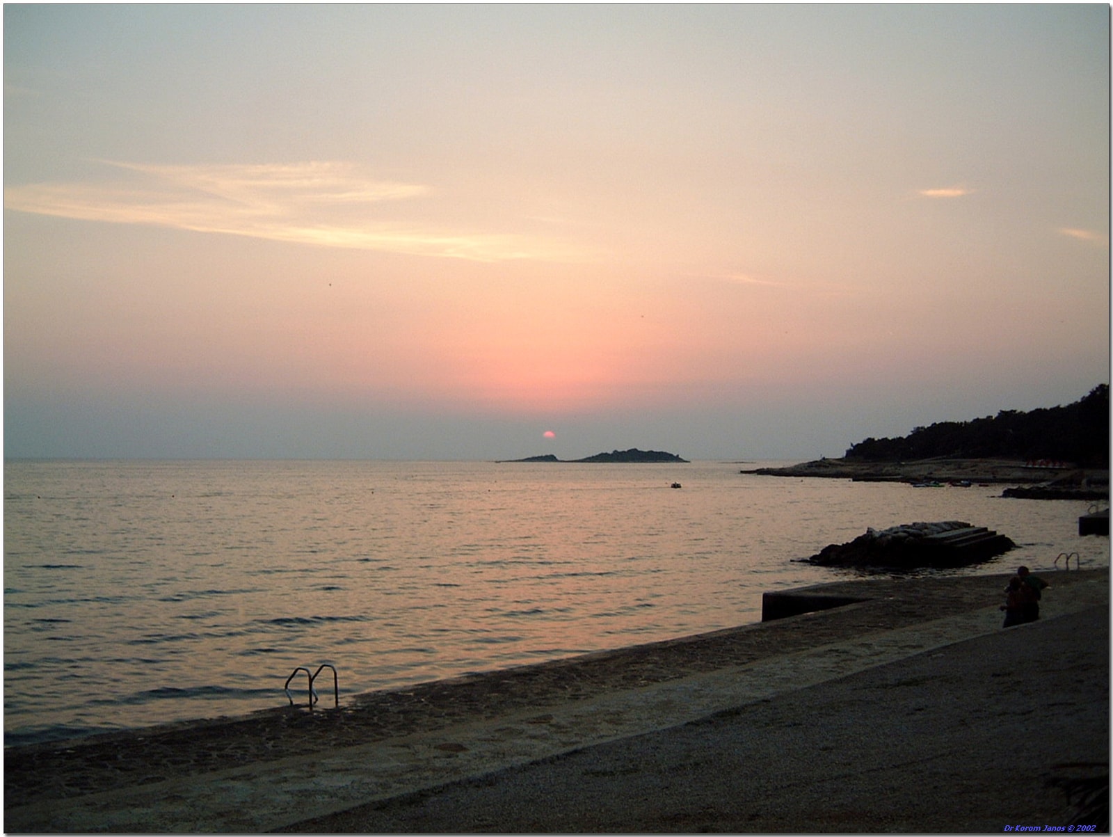 The sun sets over the calm Adriatic Sea, silhouetting a small island on the horizon, as seen from a paved beach in Poreč.