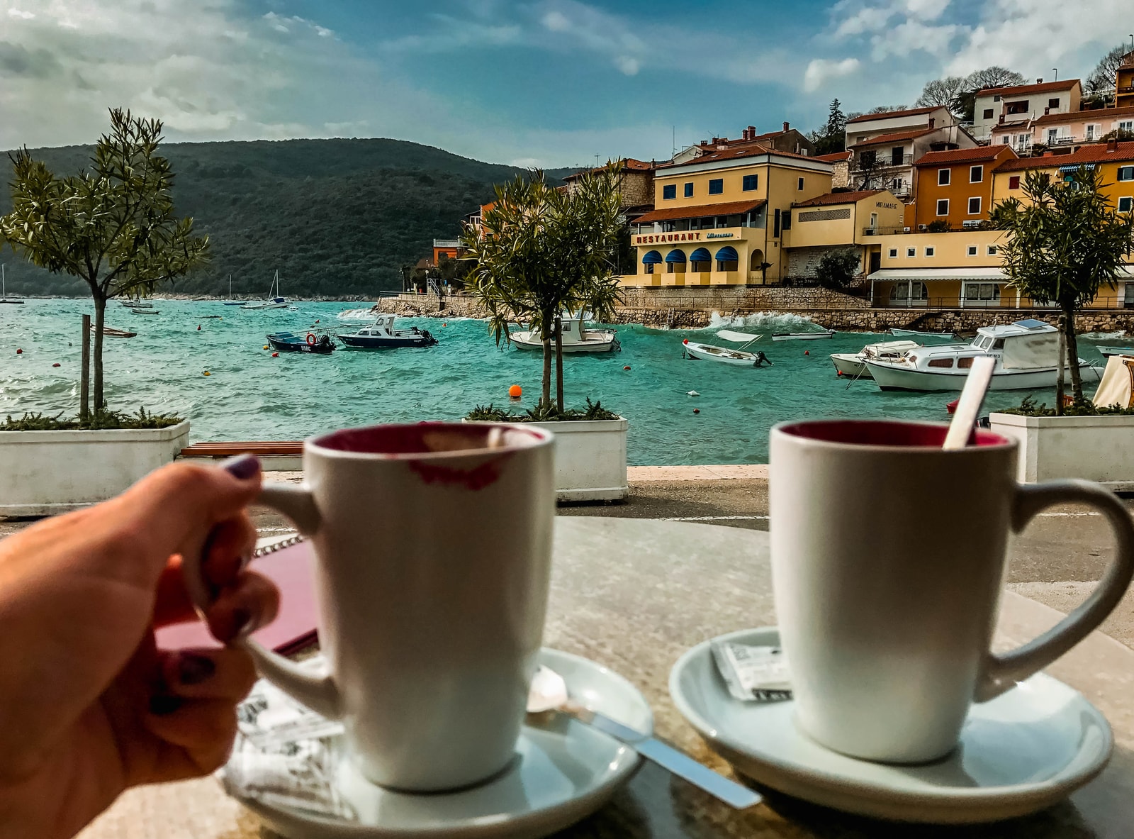 Two cups of coffee sit on a wooden table overlooking a bustling harbor beach in Rabac, capturing the relaxed seaside lifestyle.