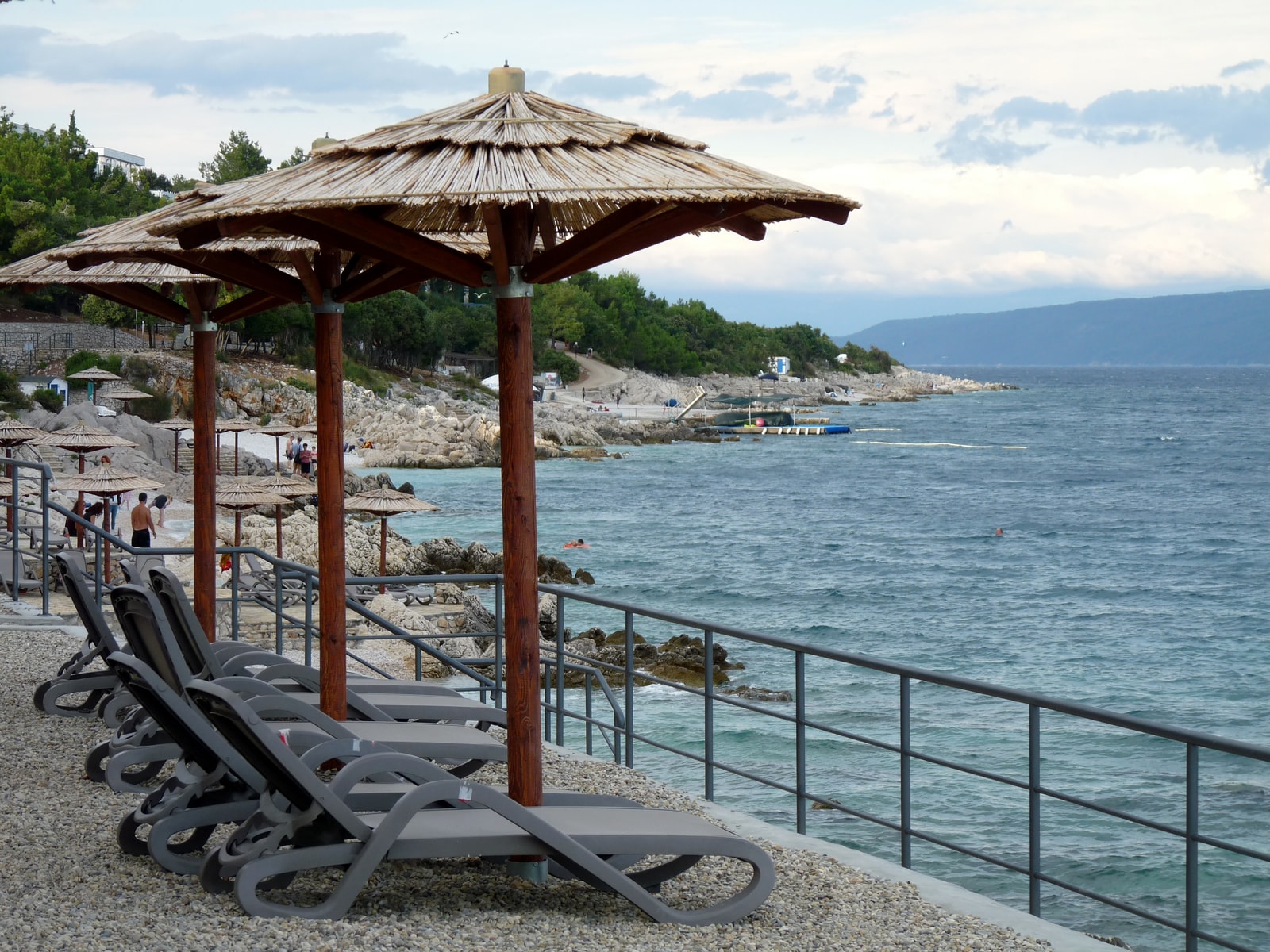 Modern white loungers with straw umbrellas line a pristine white pebble beach next to the deep blue sea in Rabac.