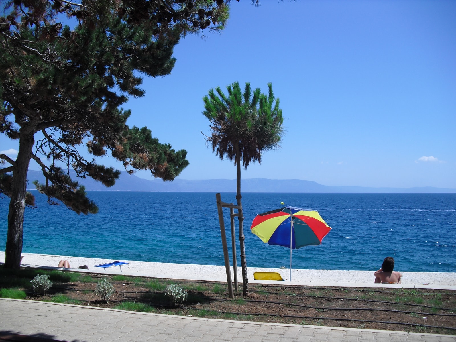 A colorful beach umbrella stands on a white pebble shore in Rabac, with the calm, deep blue sea in the background.