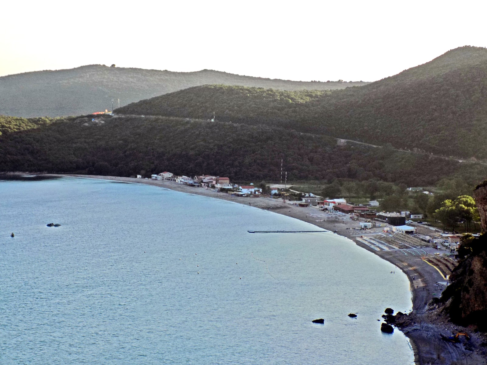 A wide pebble and sand beach with calm blue water, with a road and green hills behind it.