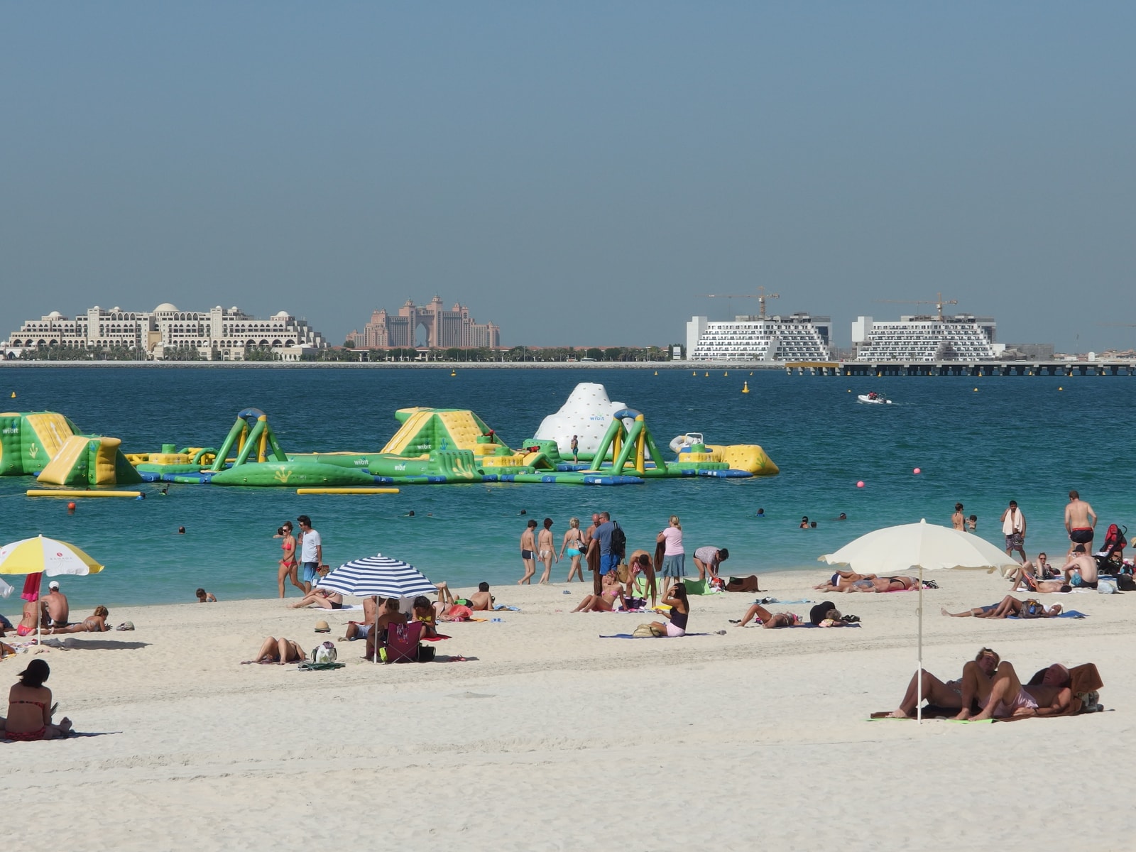 A wide sandy beach with a large inflatable water park floating in the calm blue water, with a modern city skyline in the background.