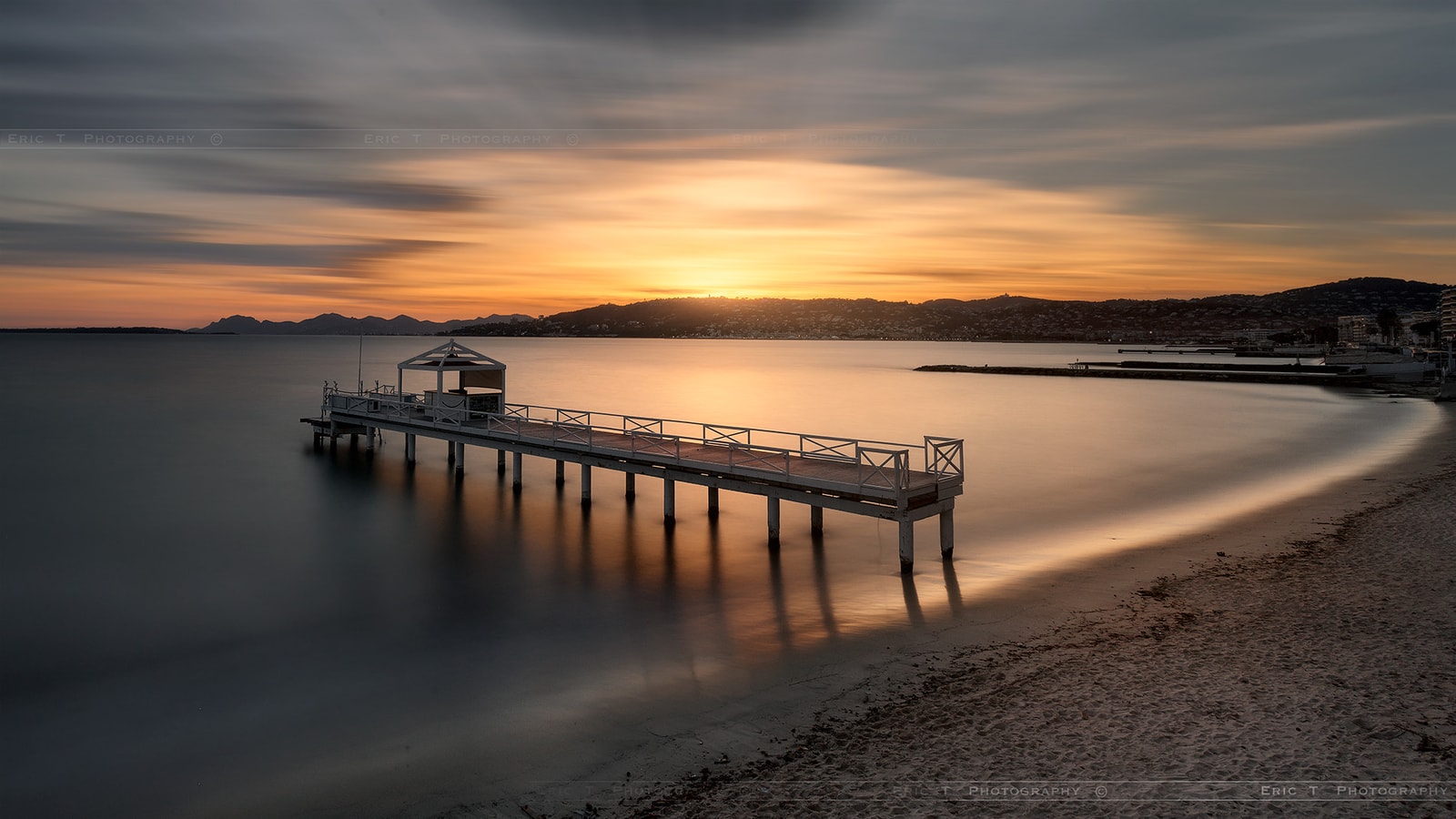 A wooden pier stretches out into the calm sea at sunset, with soft golden light reflecting on the water.
