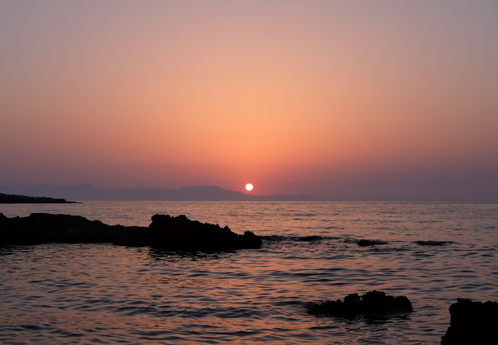 A vibrant sunset paints the sky over the calm waters and sandy shore of Kalathas Beach on the Akrotiri Peninsula.