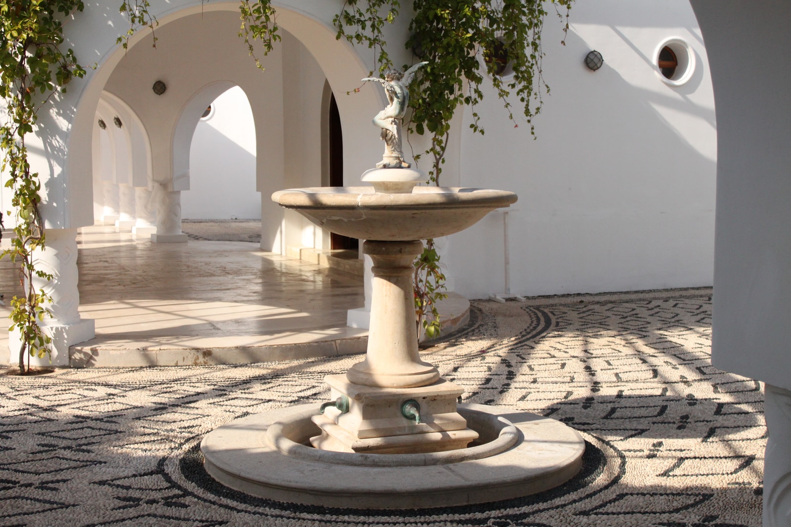 A symmetrical view of a courtyard at Kallithea Springs, featuring a central fountain, elegant arches, and patterned paving.