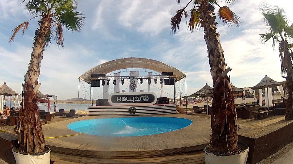 The stage and poolside cabanas of Kalypso Club, framed by tall palm trees on a summer day.