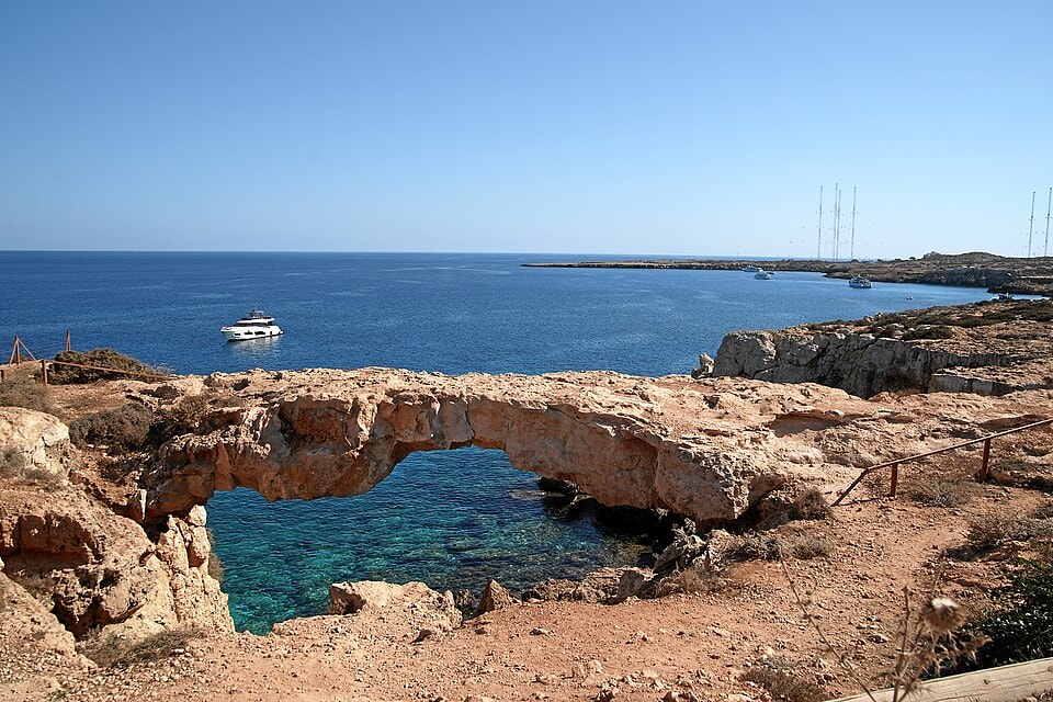 The Kamara Tou Koraka, a natural rock arch formation, stands on the coastline of Cape Greco against a blue sky.