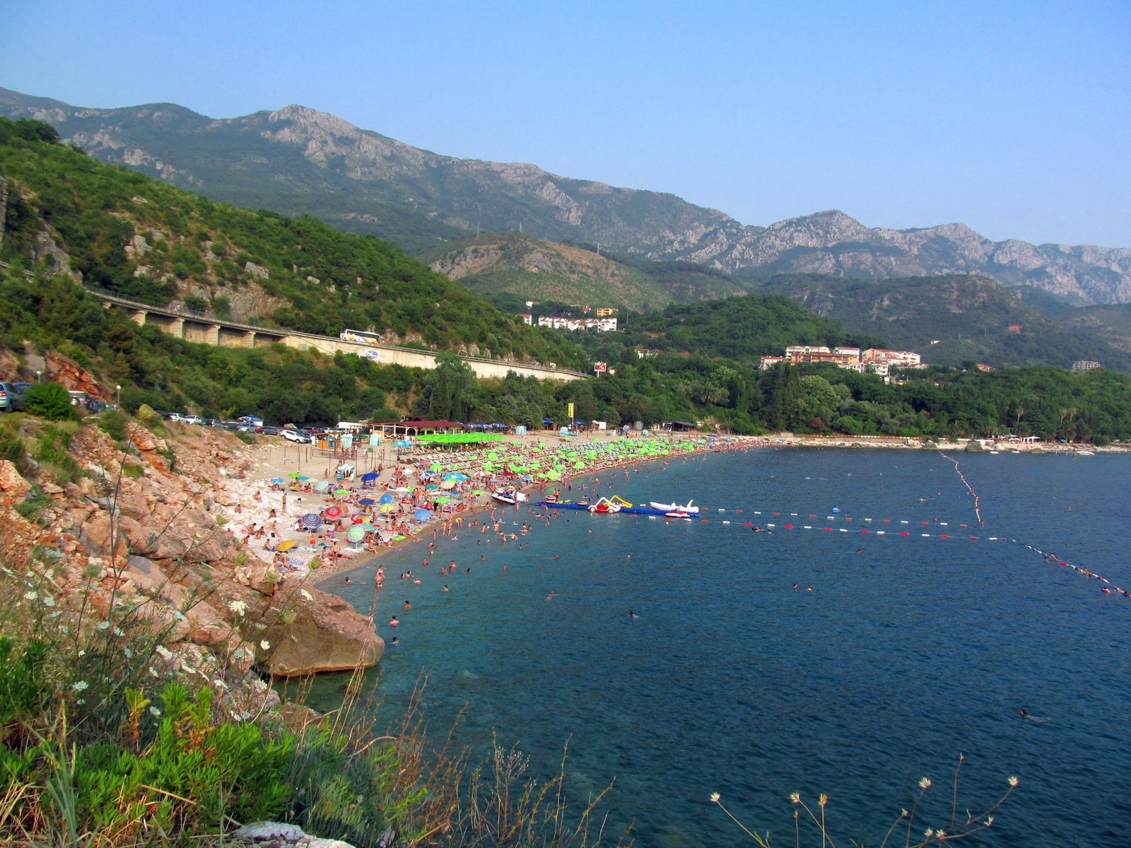 A crescent-shaped pebble beach with turquoise water is lined with umbrellas and surrounded by green hills.