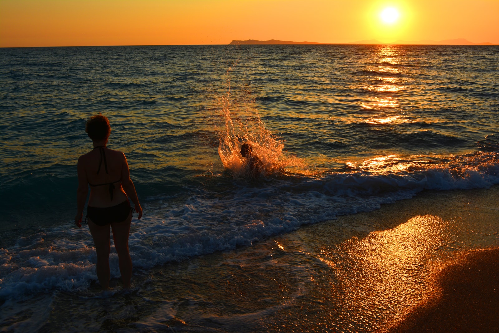 A person splashes in the water at sunset, backlit by a deep orange sun on the horizon at Karavostasi beach.