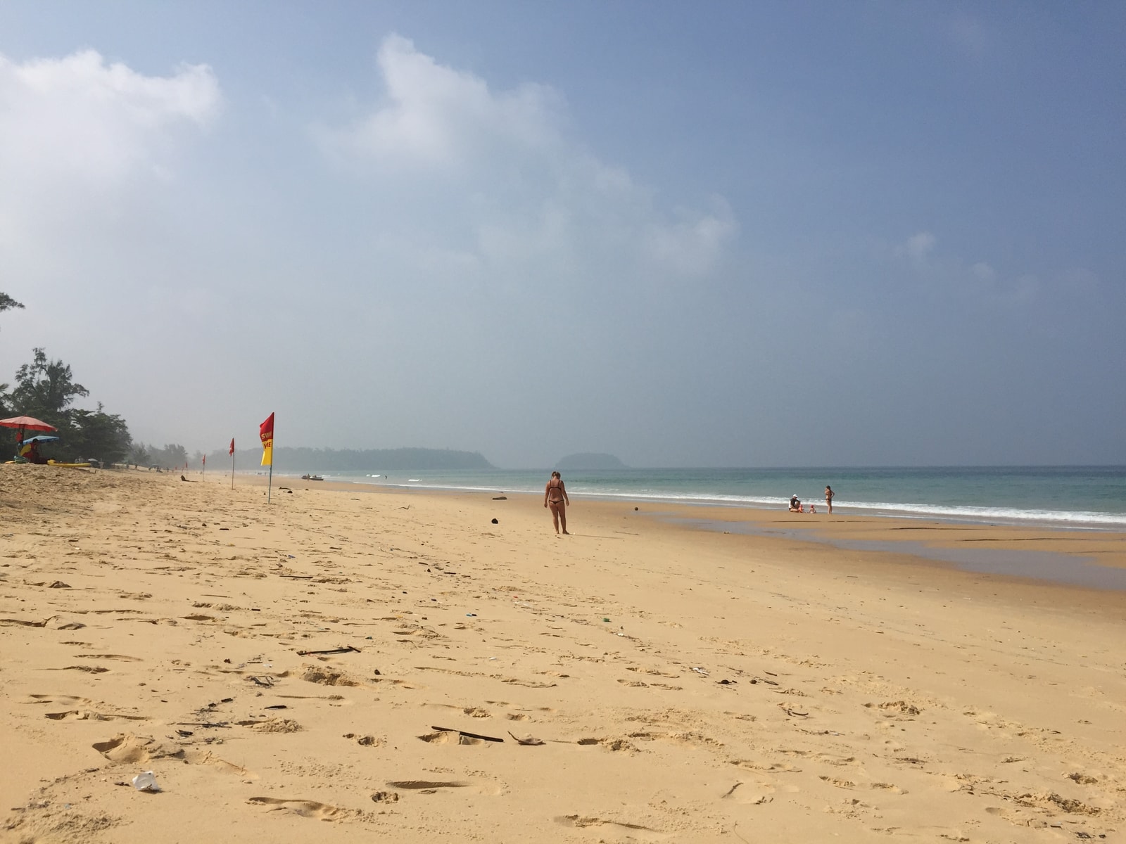A wide expanse of golden sand stretches towards the water on a clear morning, with gentle waves and a few people in the distance.
