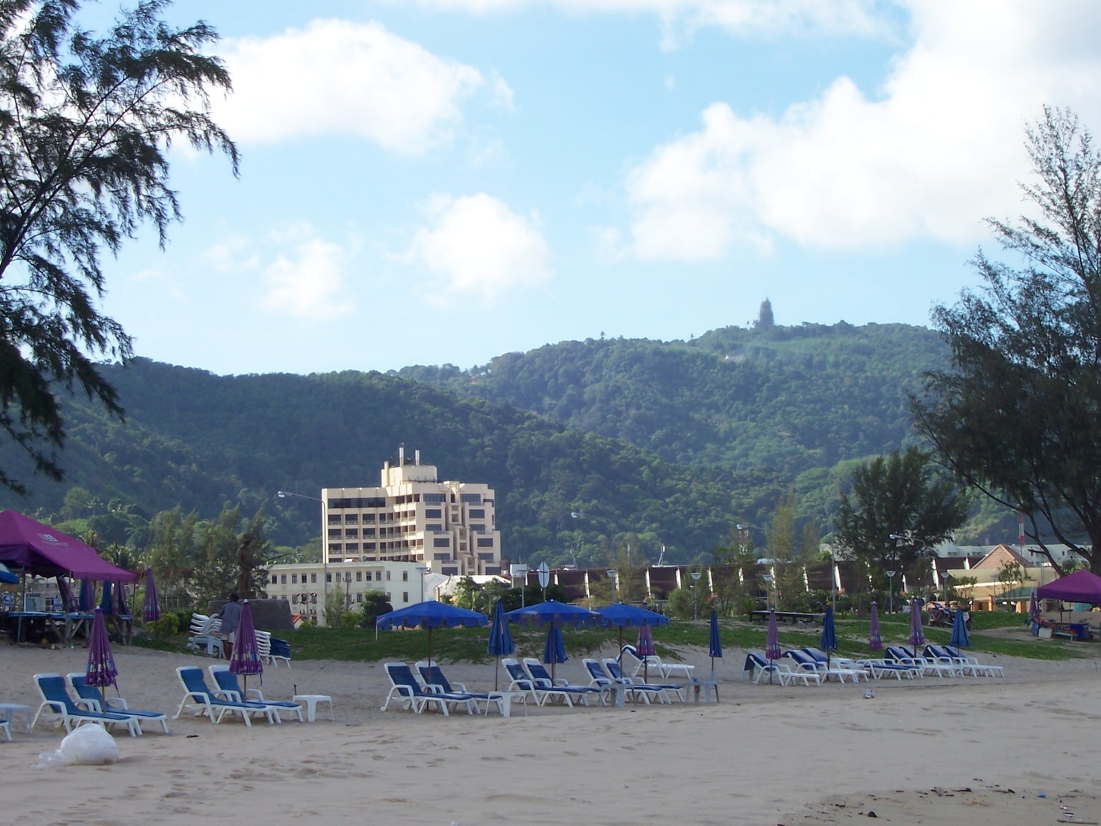 A wide sandy beach is lined with umbrellas and lounge chairs, with large resort buildings visible behind palm trees and backed by green hills.