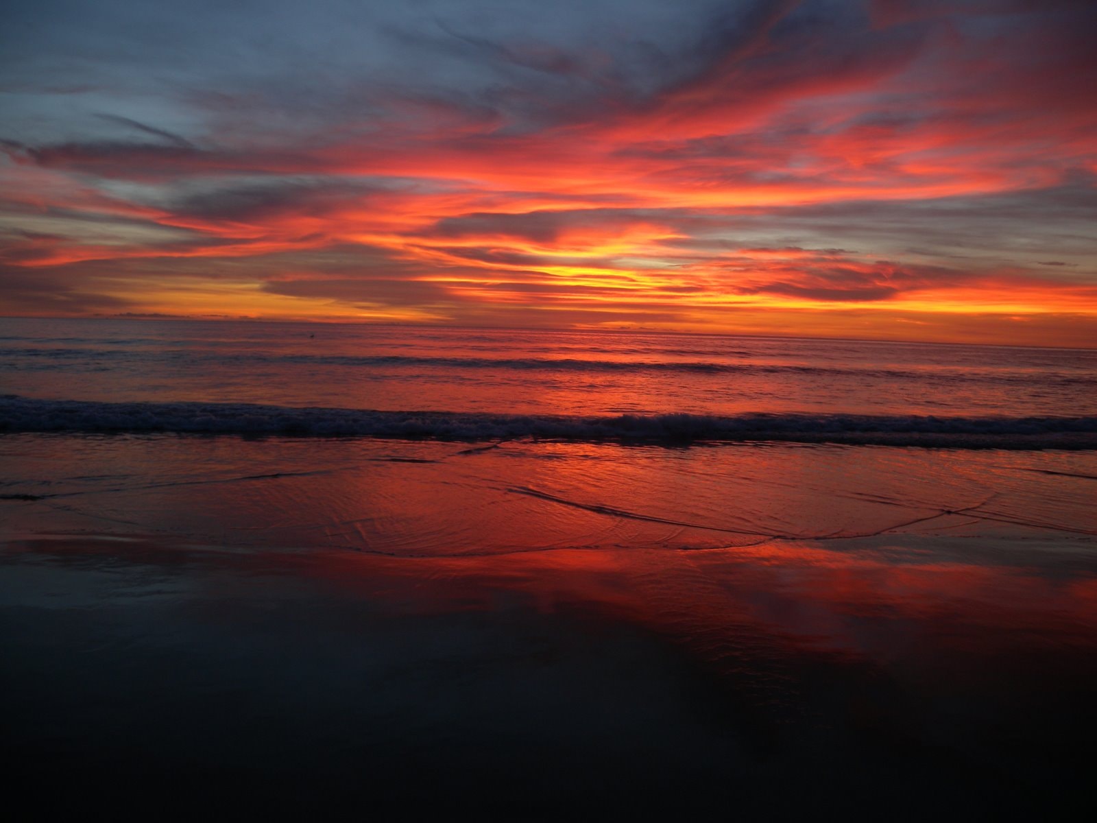 A brilliant orange and purple sunset reflects on the wet sand of a wide beach as waves gently roll in.