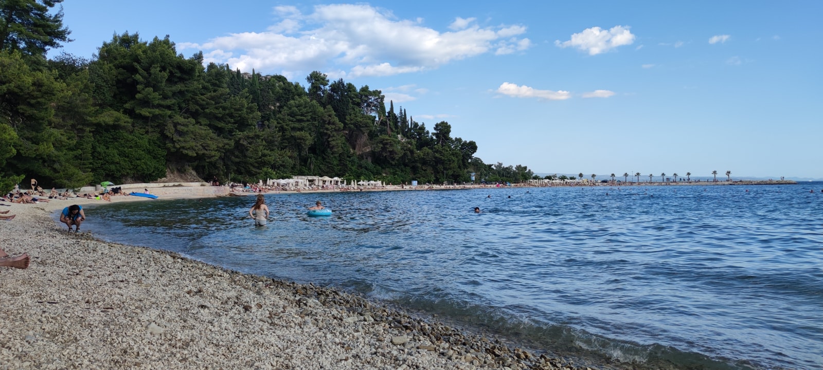 Daytime shot of Kašjuni Beach in Split, showcasing its fine pebbles and pine trees lining the coast of Marjan Hill.