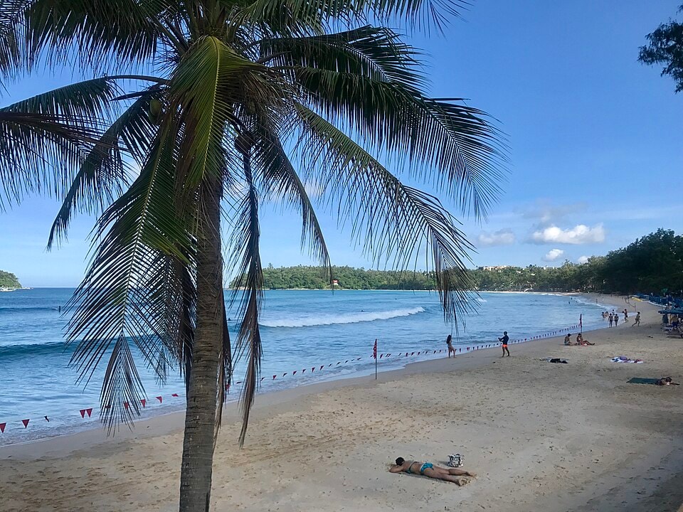 An angled view of Kata Beach shows the curve of the bay, with lush green hills rising up from the shoreline.