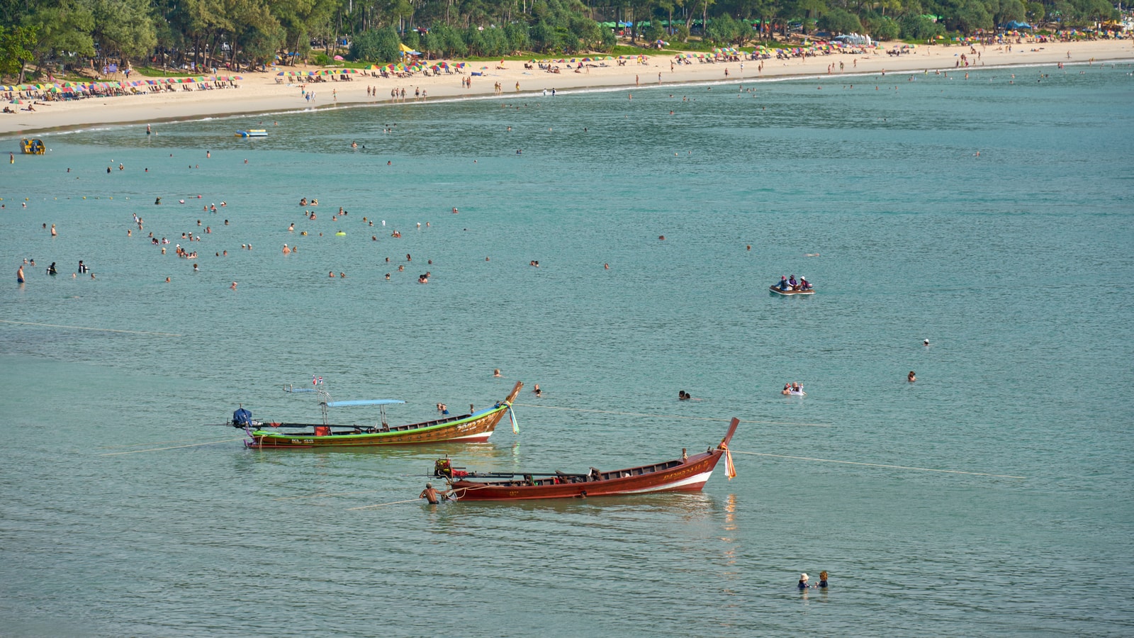 Longtail boats are moored in the calm, turquoise water of a wide bay, with a sandy beach and lush green hills in the background.