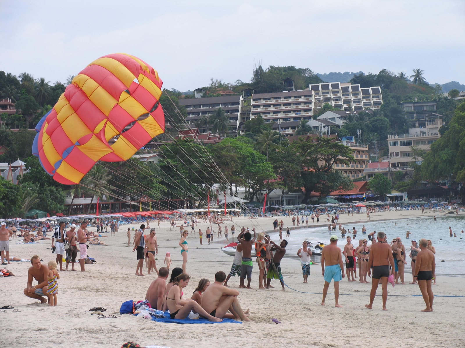 A person parasailing high above the water at Kata Beach, with the resort-backed shoreline visible below.