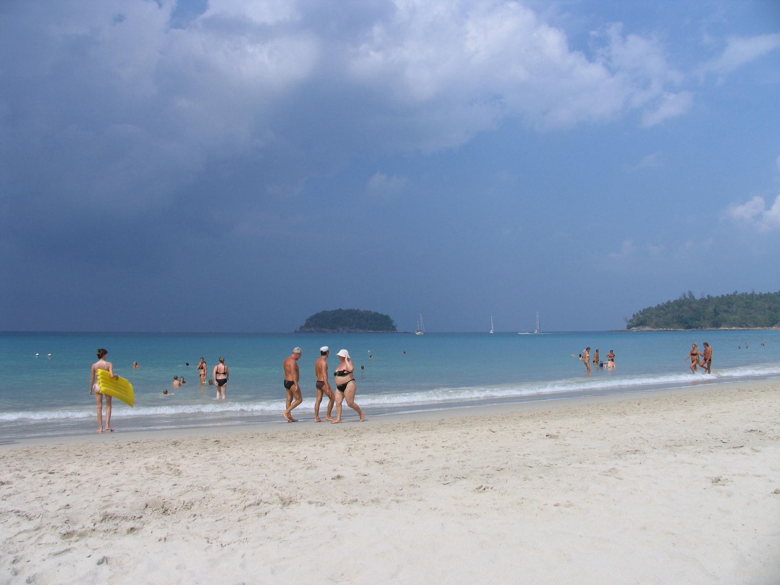 Dark storm clouds gather over Kata Beach, with people still visible on the sand and in the water.