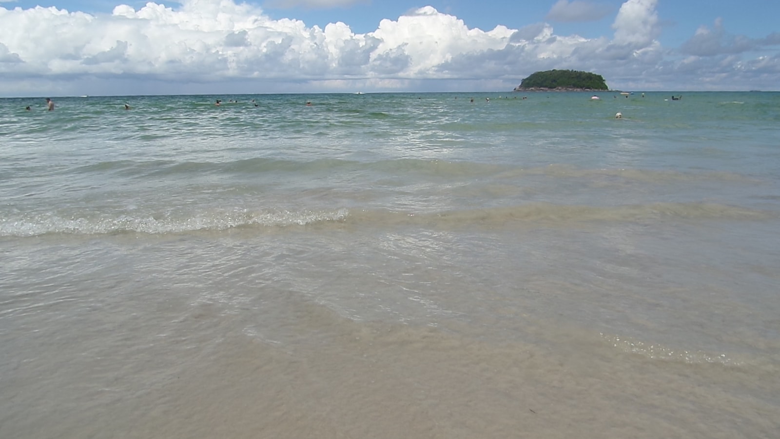 A calm blue sea gently laps a sandy shore under a bright, sunny sky, with a small island visible in the distance.