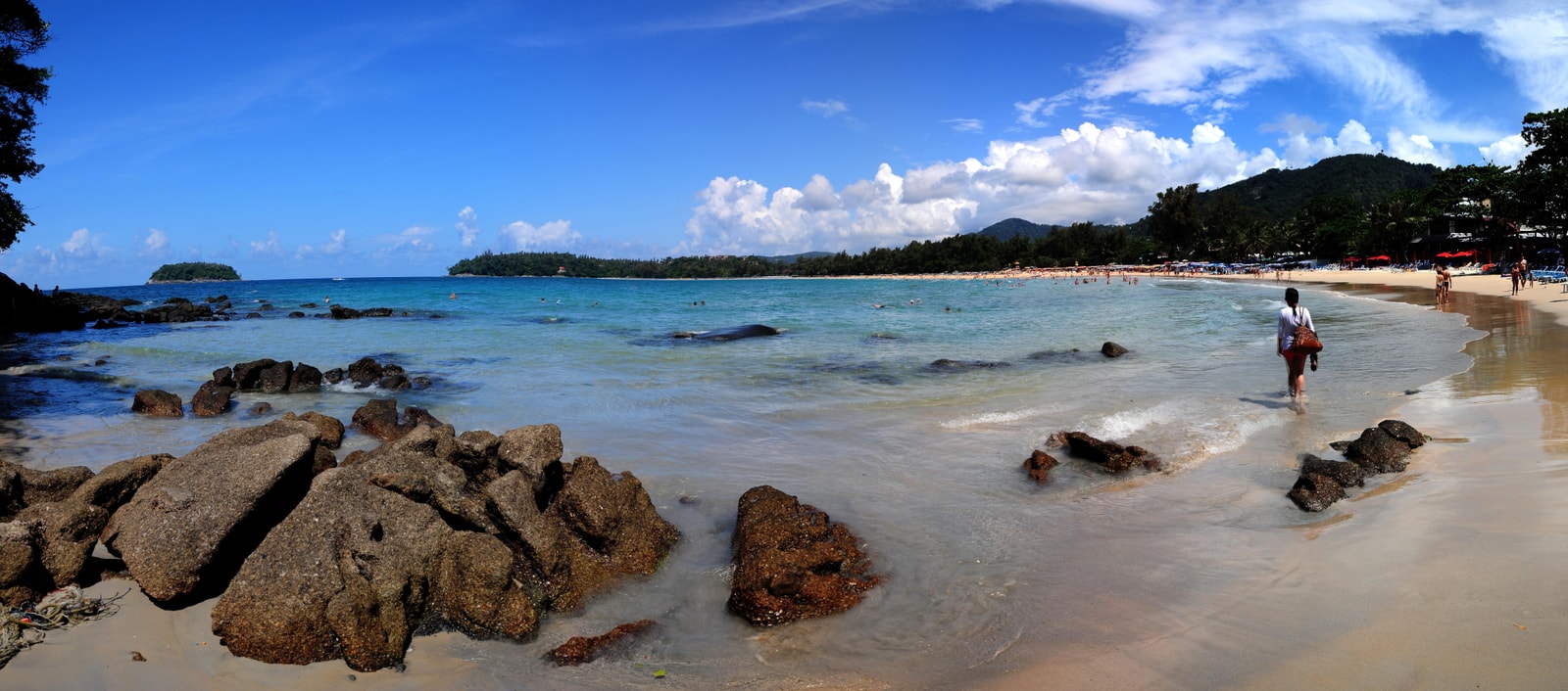 A wide panoramic shot of Kata Beach showing a rocky foreground, the sandy bay, and a small island offshore.