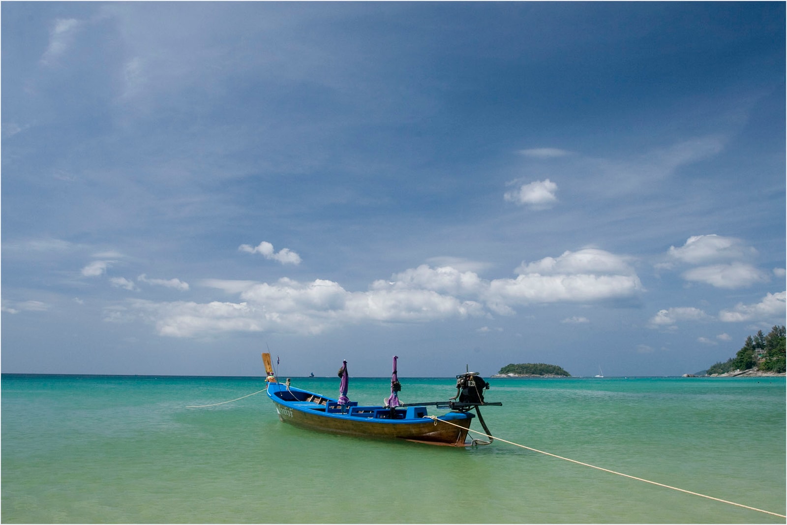 A traditional long-tail boat with colorful ribbons is moored in the calm, clear water of Kata Noi beach.