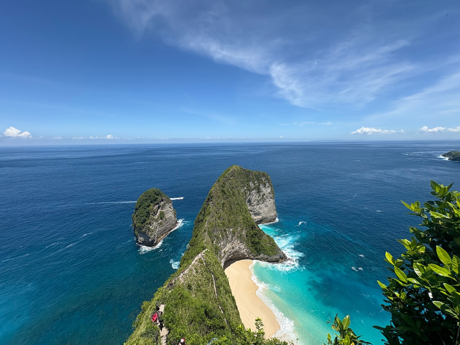 A view of the Kelingking promontory, with its white sand beach, turquoise shallows, and breaking surf in the ocean.