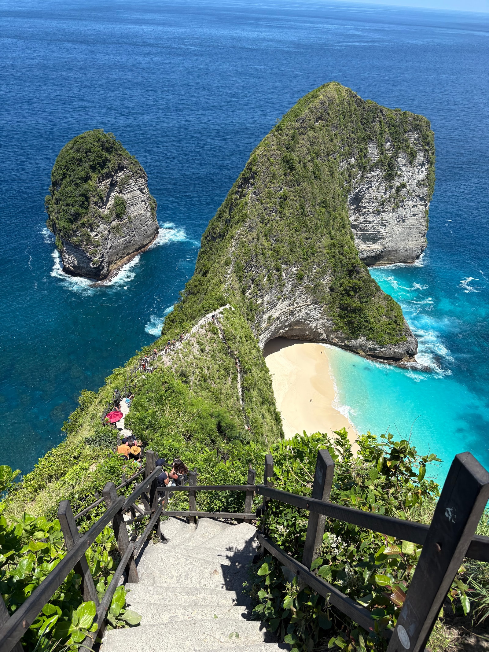 A vertical shot shows the steep stairs and path leading down the cliffside ridge at Kelingking Beach, with the ocean far below.