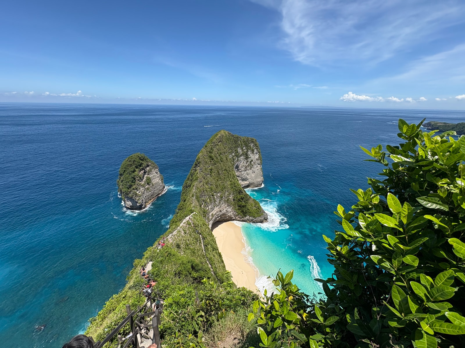A wide view from the Kelingking viewpoint shows the horizon, distant mountains, and the full expanse of the bay with visitors for scale on the cliff edge.