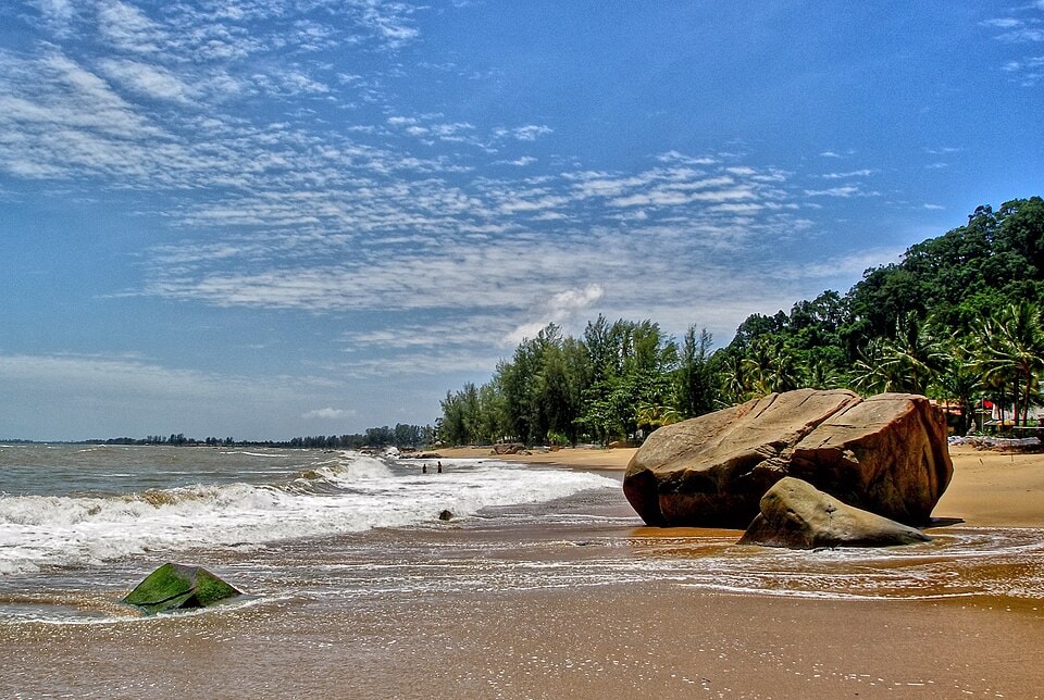 View of Khao Lak Beach with large boulders in the foreground, foamy surf, and coastal vegetation under a vivid sky.