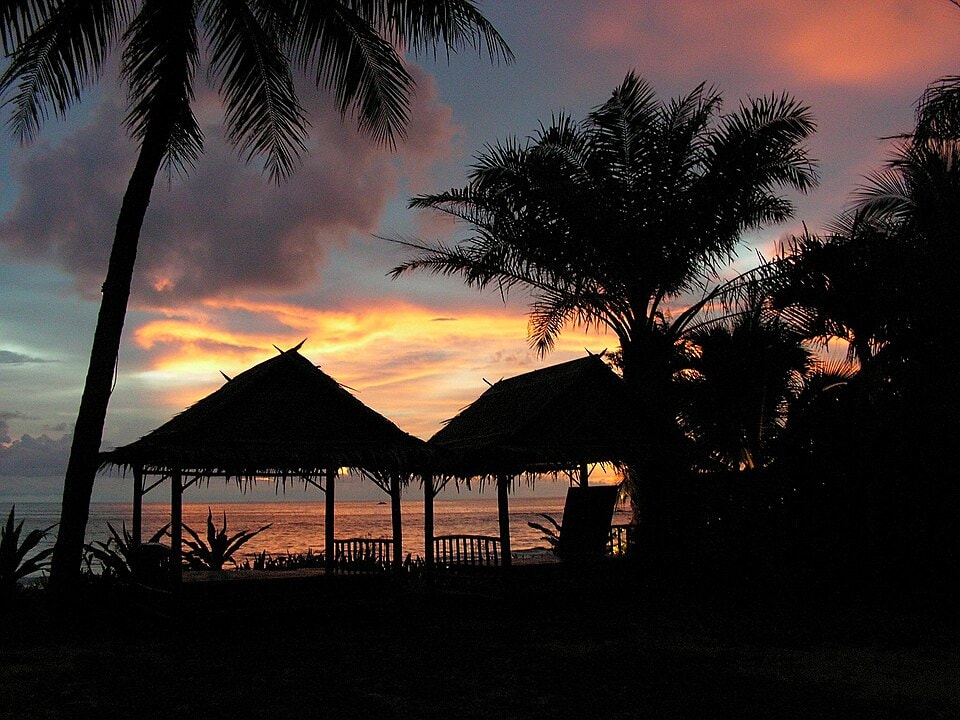 A colorful sunset over the water in Khao Lak, with palm tree silhouettes and waterfront huts.