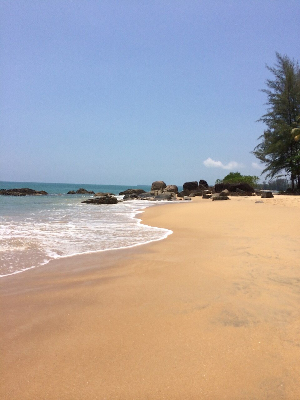 A peaceful view of Khuk Khak Beach showing calm, shallow surf, a sandy shore with some rocks, and a line of coastal trees.