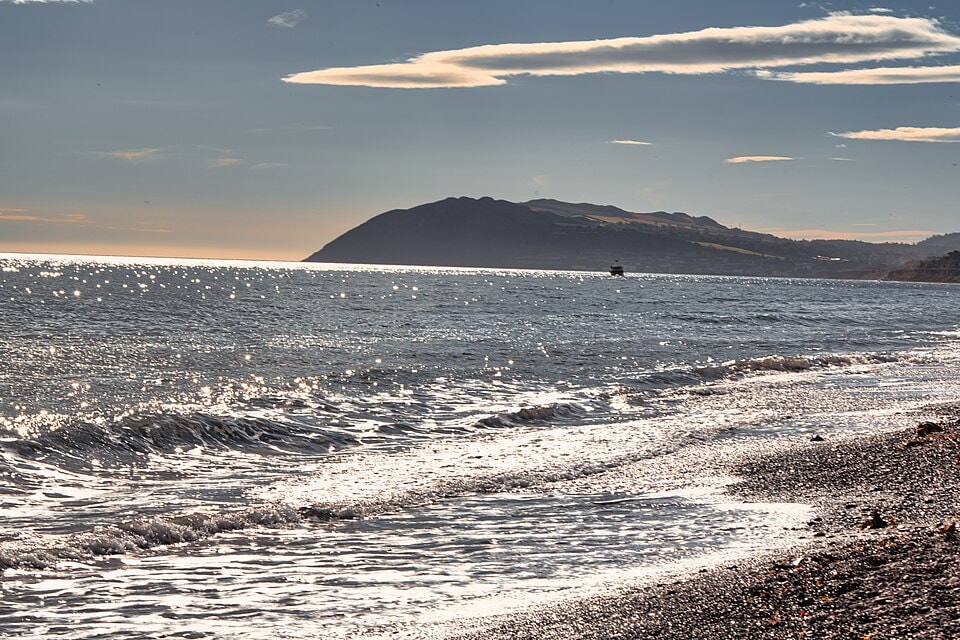 Backlit waves crash on a beach with a dark headland in the background, creating a dramatic, warm-toned scene.