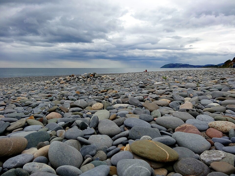 A pebbled beach with dramatic clouds overhead and a headland in the distance.