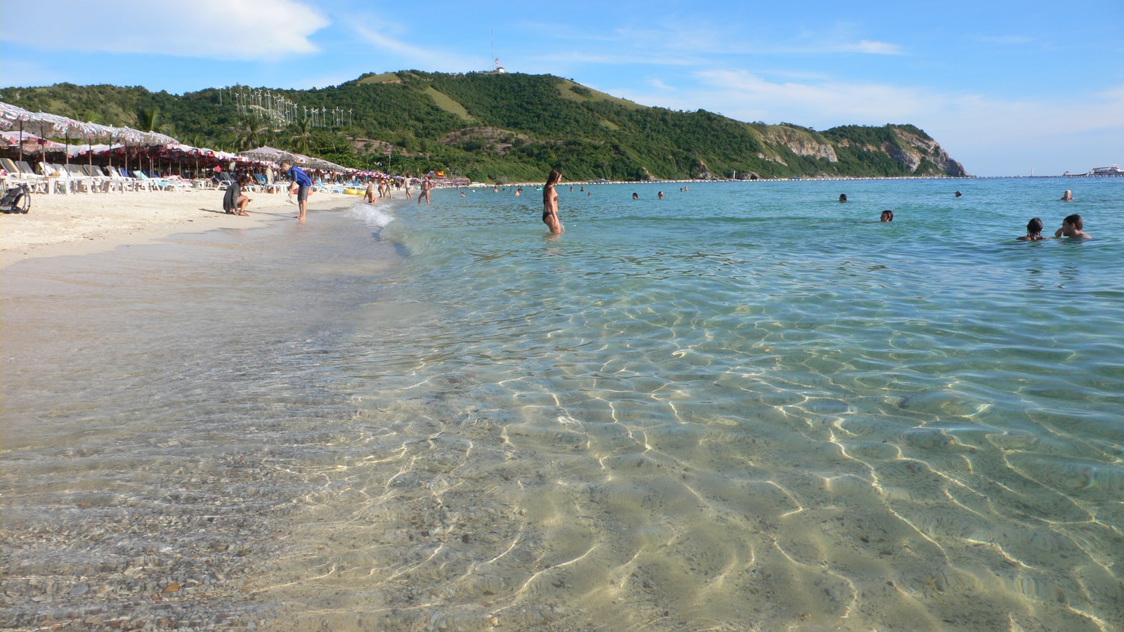 A view of a curved beach on Koh Larn with clear shallow water, green hills, and resort buildings.