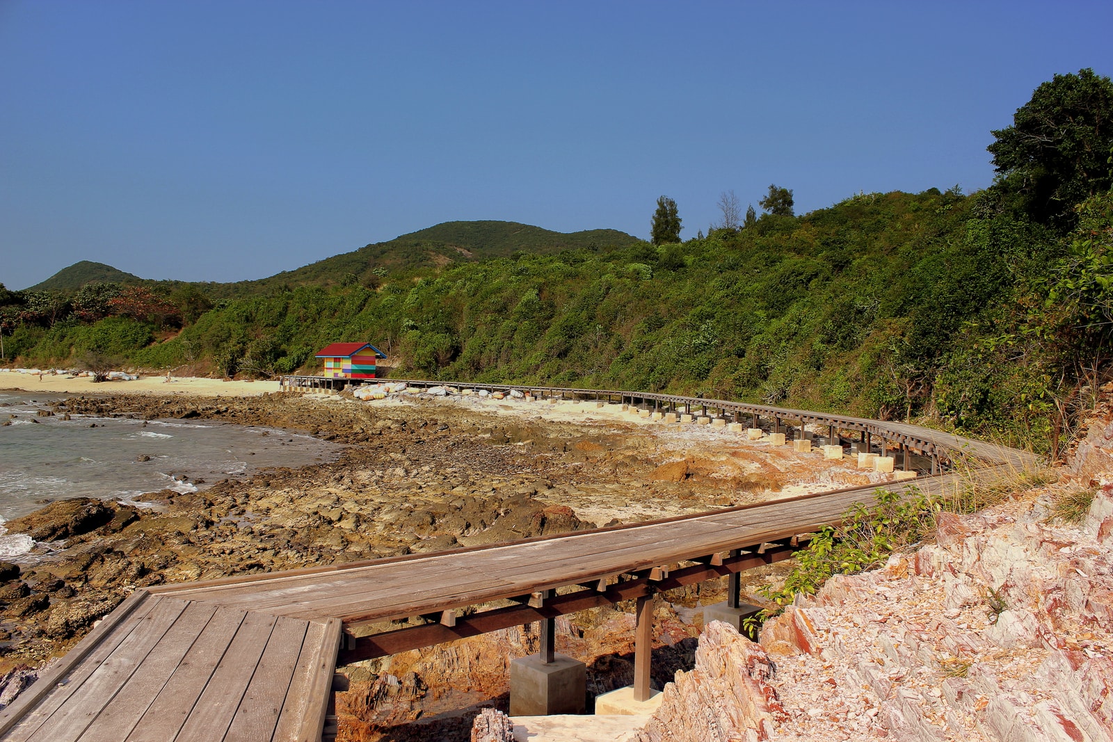 A wooden boardwalk winds along a rocky coastline on Koh Larn, with lush green vegetation on the hillside.