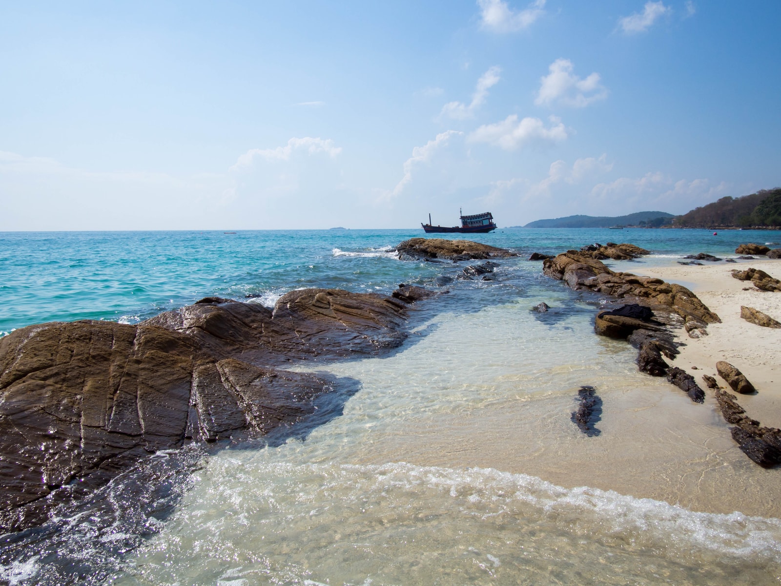 A rocky inlet on Koh Samet with calm, shallow turquoise water, white sand, and a small boat moored near the shore.