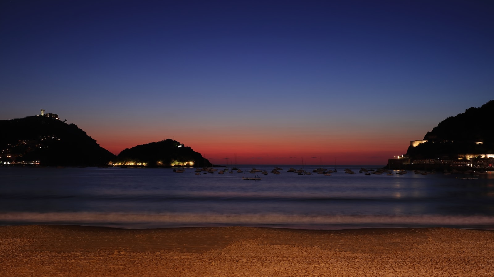 A long-exposure shot of La Concha Bay at twilight, with the water appearing silky smooth under an indigo and orange sky and the silhouettes of the island and headlands.