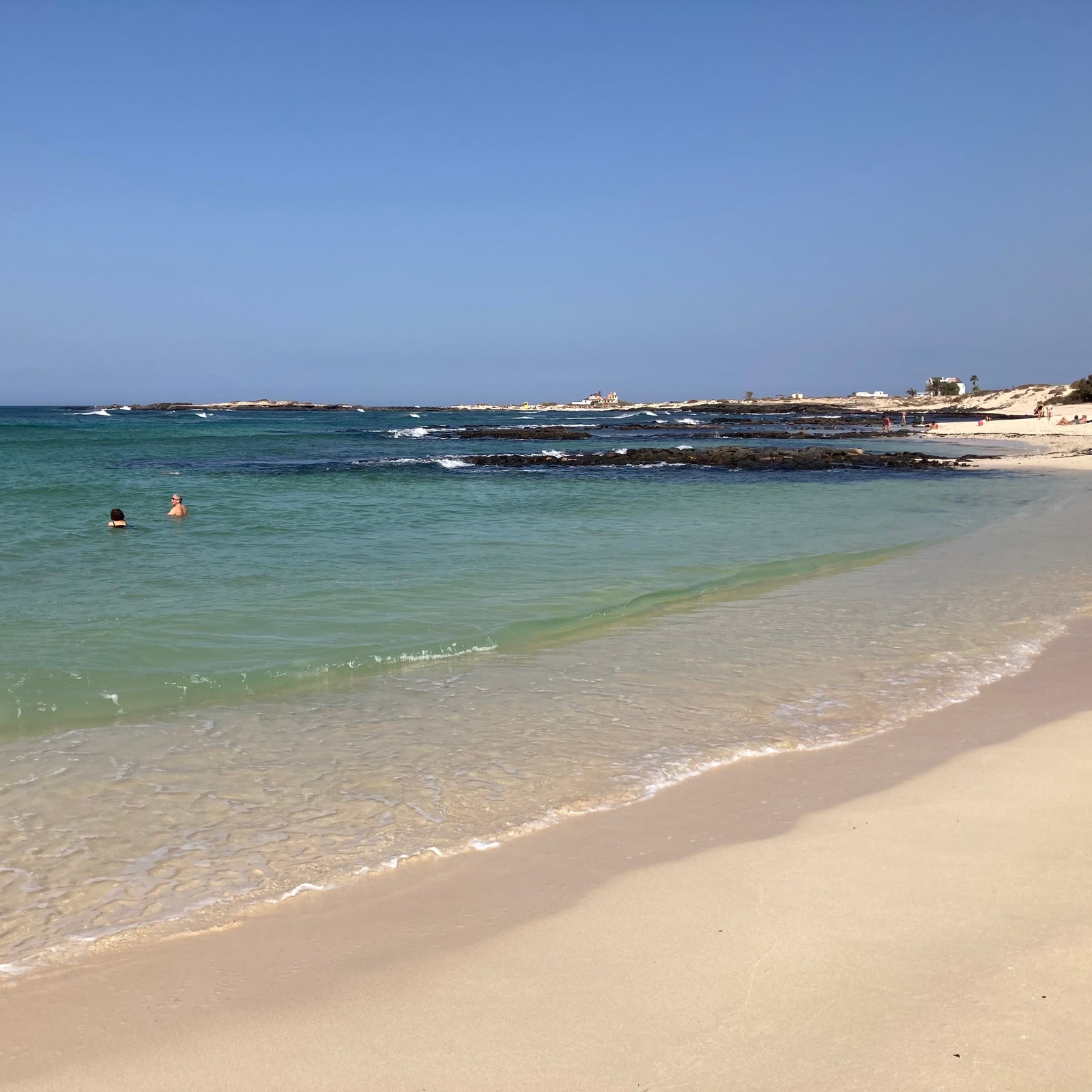 Shallow, clear green water laps against a sandy shore, with dark volcanic rocks forming a natural breakwater.