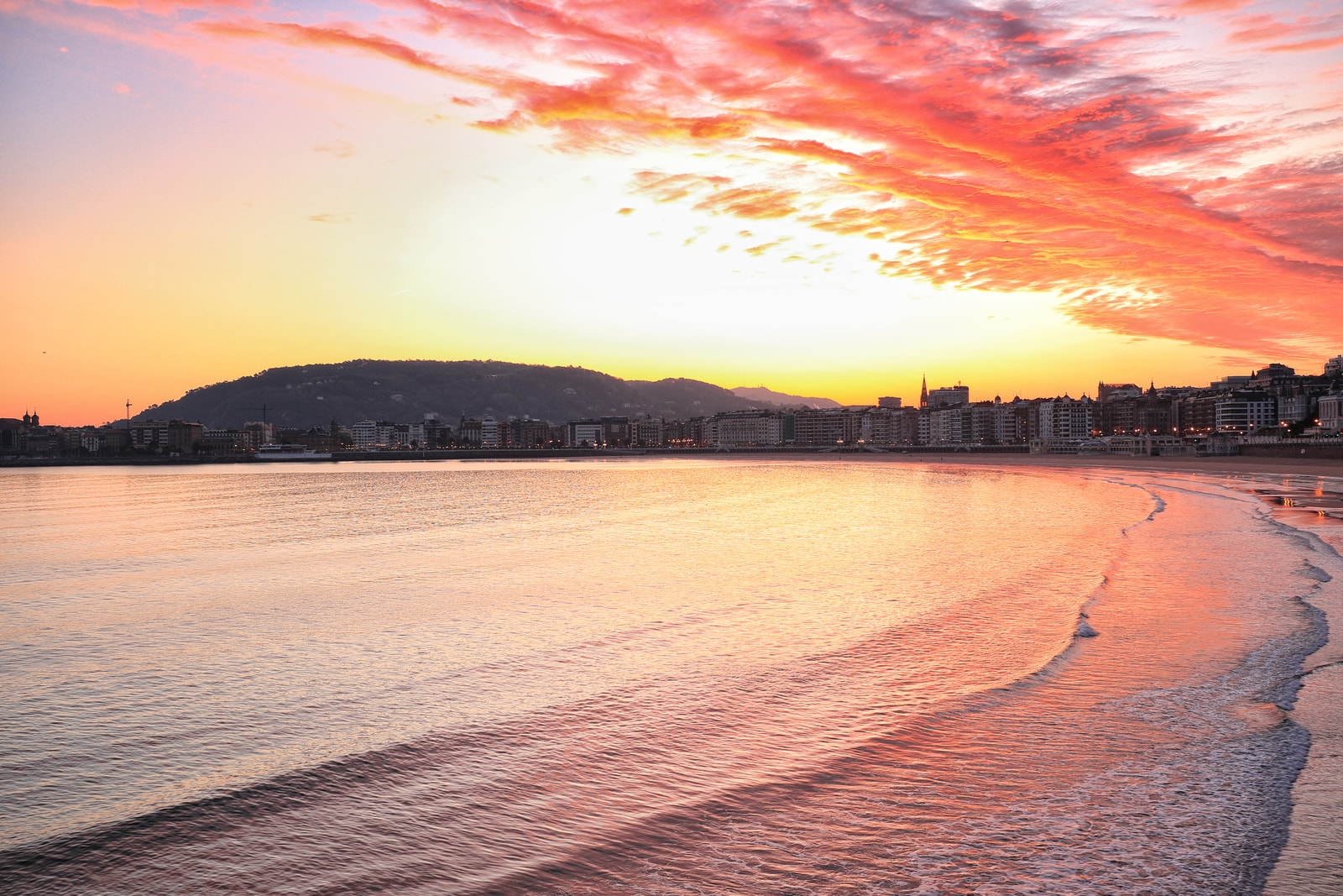A fiery sunrise over La Concha Beach, with vibrant orange and pink clouds reflecting on the smooth, wet sand during low tide.