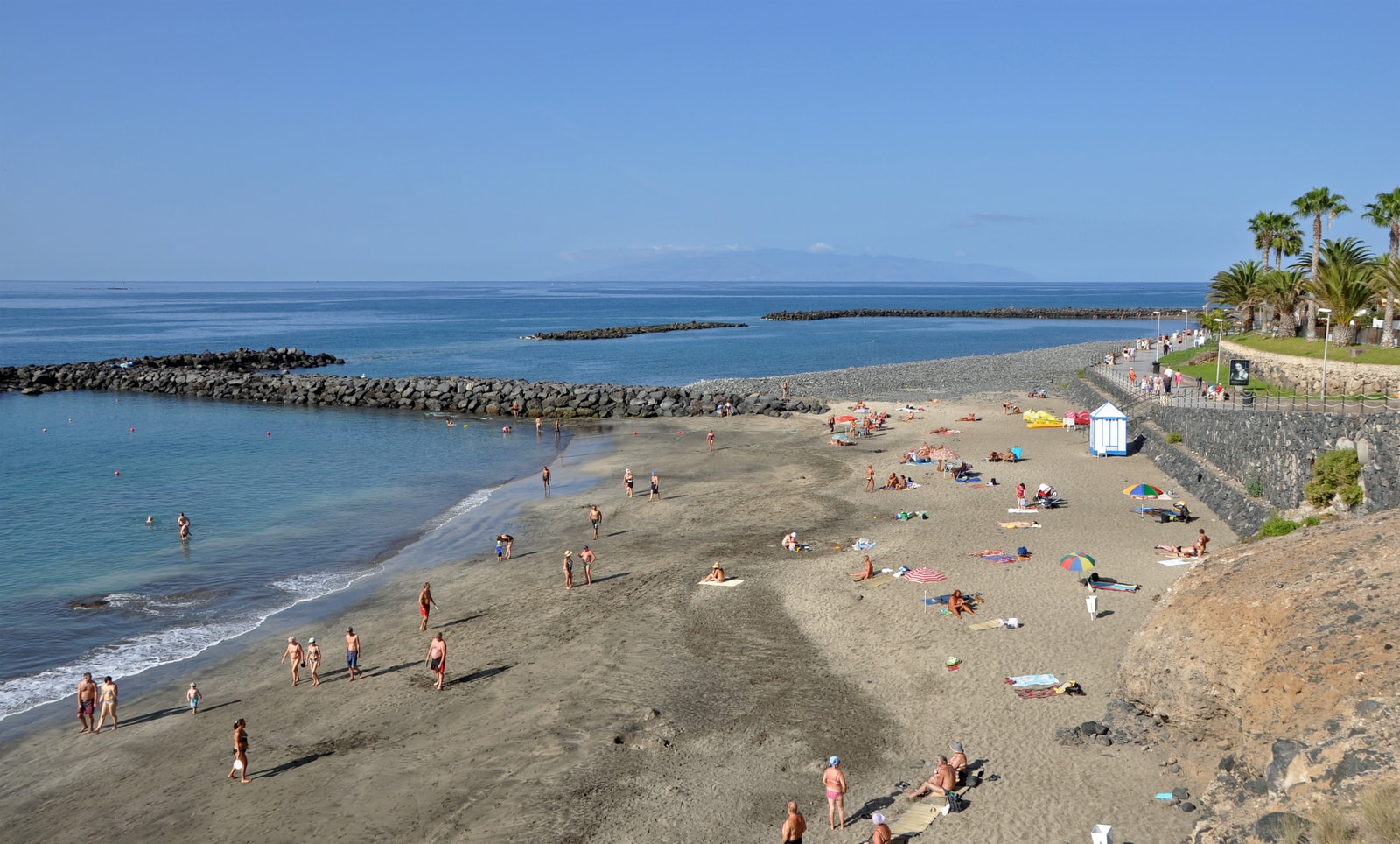 An elevated view of a beach with dark sand and pebbles, bordered by volcanic rock breakwaters.