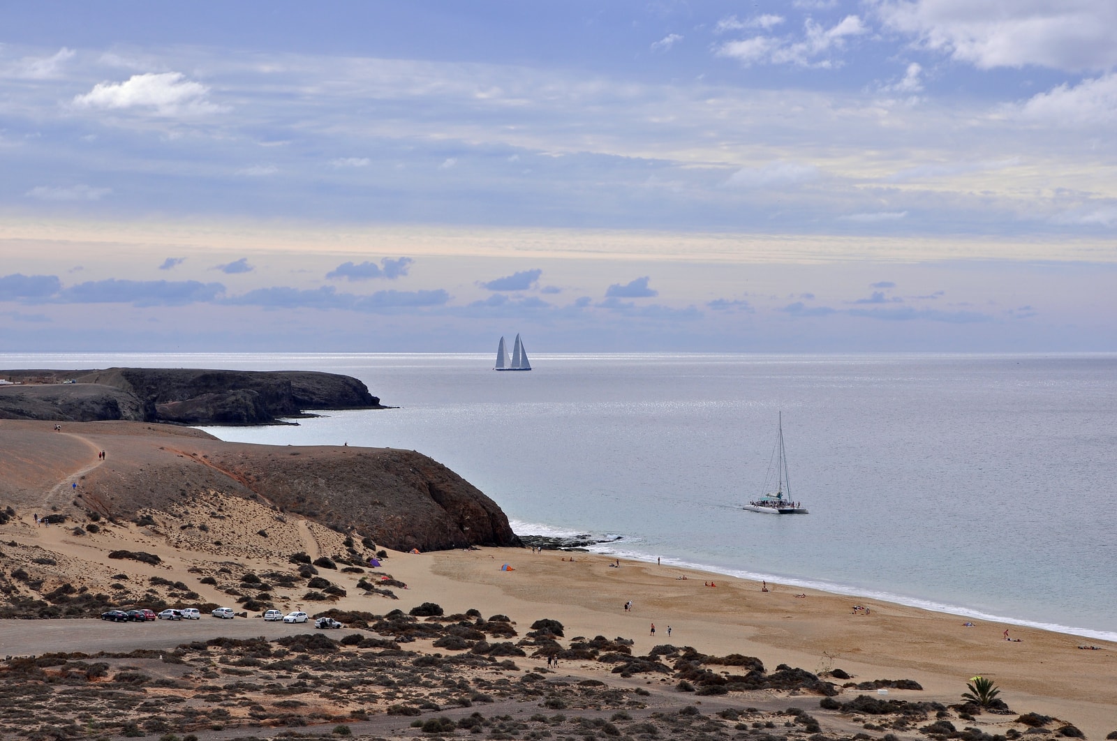 An elevated view shows a sandy cove with several boats anchored in the turquoise water, flanked by rugged, rocky cliffs.
