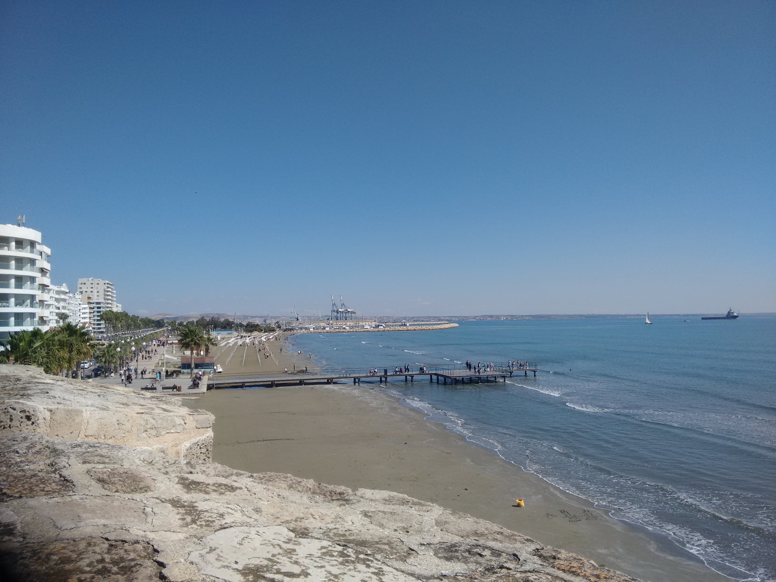 An elevated view of Larnaca's coastline, showing the beach, promenade, hotels, and a harbour with boats.