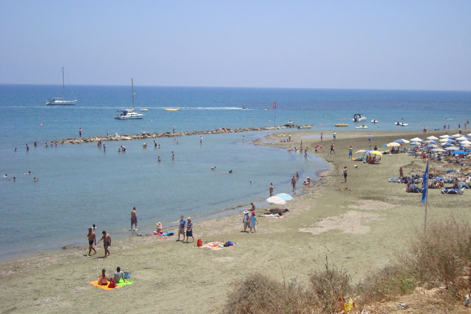 A view of Faros Beach in Pervolia, showing calm, shallow water with swimmers and beach umbrellas.